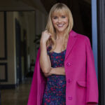 Reese stands in a doorway wearing a blue and pink print dress and a bright pink coat